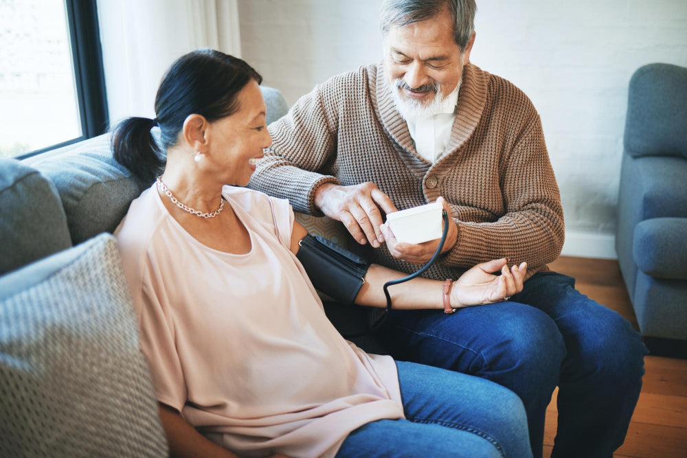 An older couple checking each other's blood pressure at home, showing everyday caregiver support in a shared living space.
