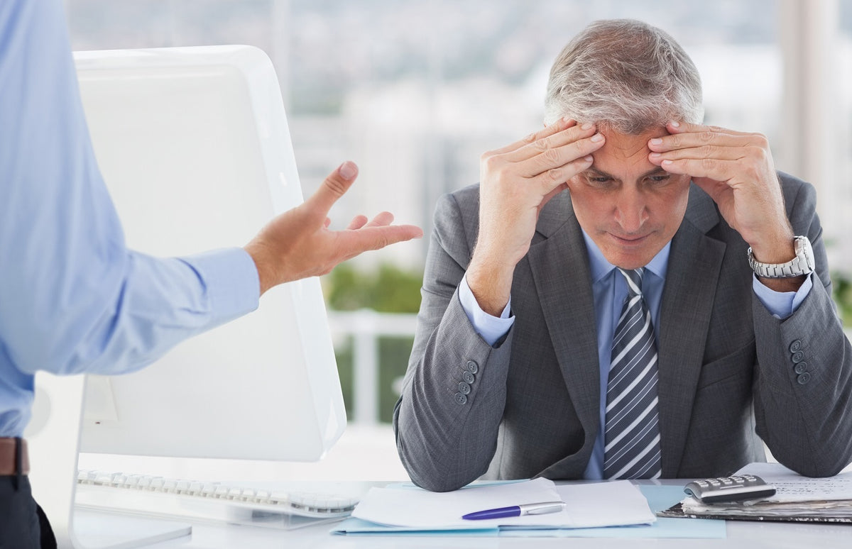 A frustrated businessman in a suit holding his forehead while another person gestures during a discussion at the office.