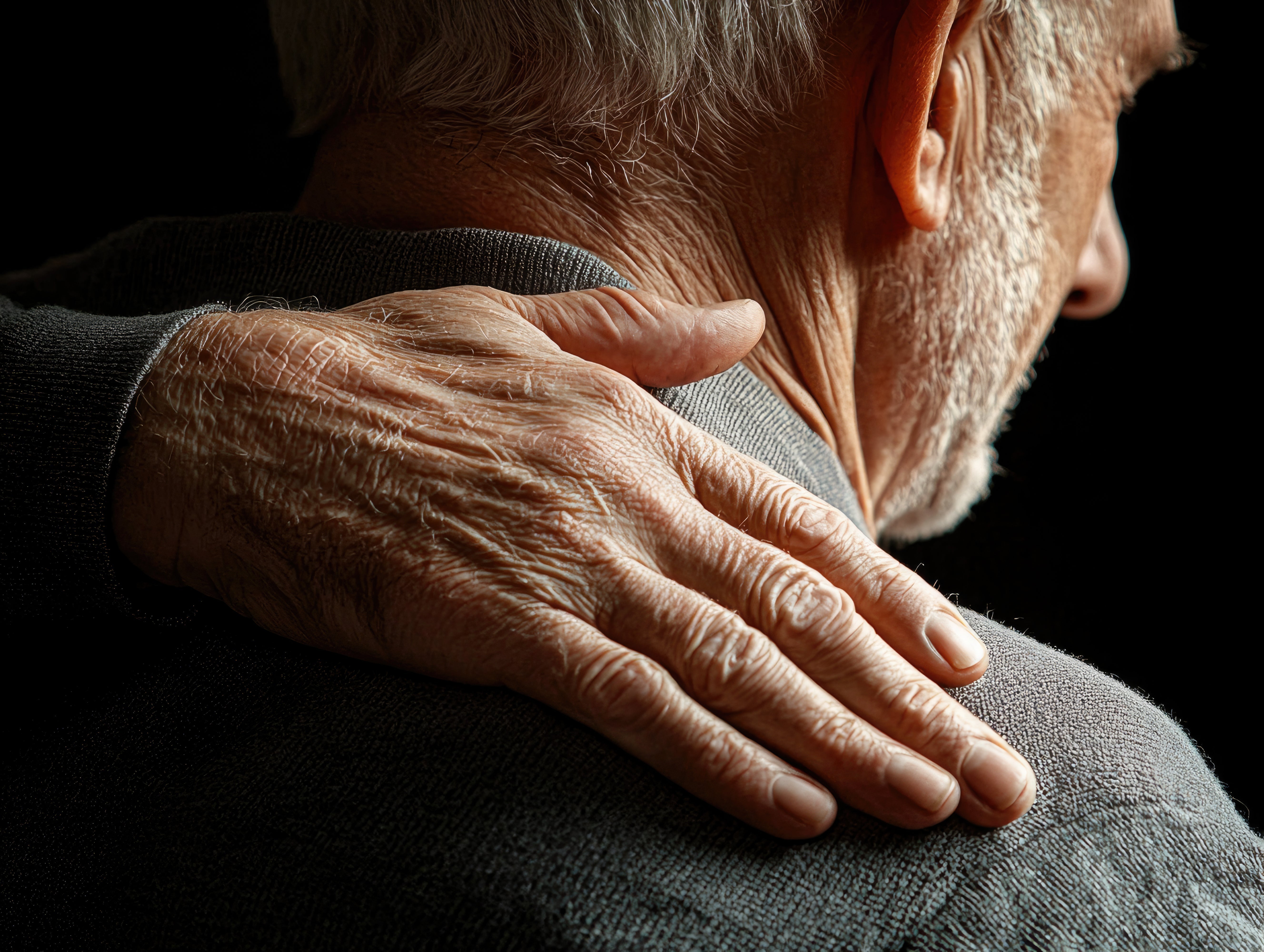 Elderly person resting hand on shoulder in low light, reflecting hereditary Parkinson's Disease family history concerns.
