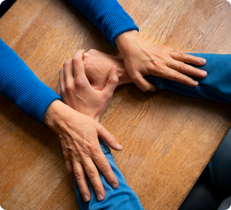 Two people at a wooden table with one person placing both hands over another's forearm in a supportive gesture, representing tremor support at work.