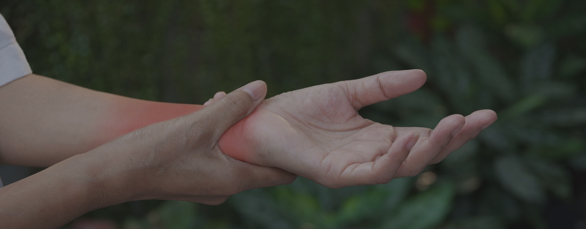A close-up of a person's hand holding another hand's wrist, with a red heat-like glow indicating pain or discomfort, set against a blurred green outdoor background.