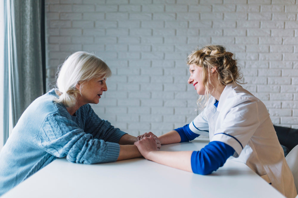 Side view of a woman talking to a nurse in a nursing home while holding hands with her.