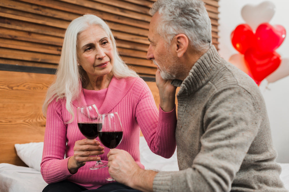 Older couple sharing red wine in a romantic bedroom setting, the woman holding two glasses and tenderly touching her partner's face.