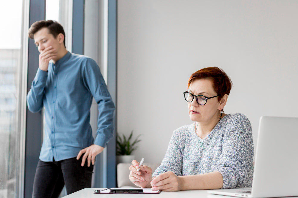 Young man standing anxiously by a window while a woman seated at a desk with a laptop takes notes, depicting a stressful workplace disclosure meeting.