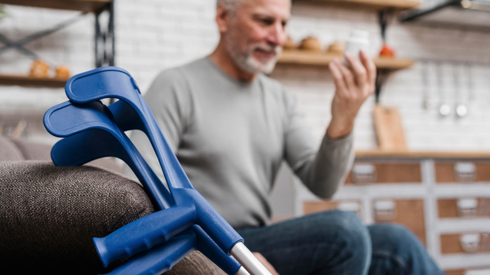 Older adult with hand tremor examining an object while seated at home, with forearm crutches in foreground highlighting mobility support and daily challenges.