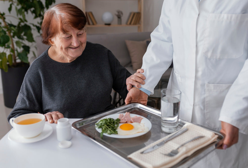 Caregiver assisting older adult with meal tray and steady support, illustrating adaptive care and daily living support for Parkinson’s patients.