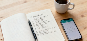 An open notebook with handwritten questions beside a coffee cup and a phone showing a calendar, planning what to ask at a medical appointment.