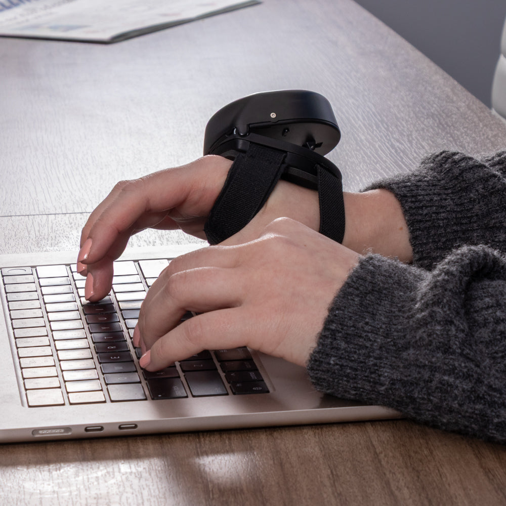 Close-up of hands typing on a laptop, with the Steadi-3 anti-tremor device secured on the back of one hand with a black strap while working at a desk.