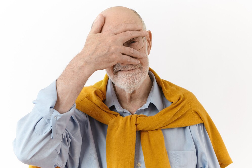 An Elderly man playfully covering one eye with one hand, wearing glasses, a light blue shirt, and a mustard yellow sweater.